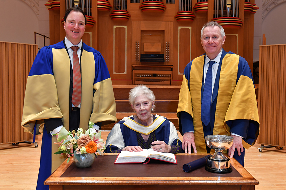 A woman with short grey hair sat signing a book in front of an organ. Two men stand either side wearing robes.